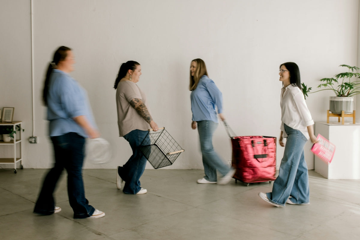 Four women walk across a minimalist white room with belongings